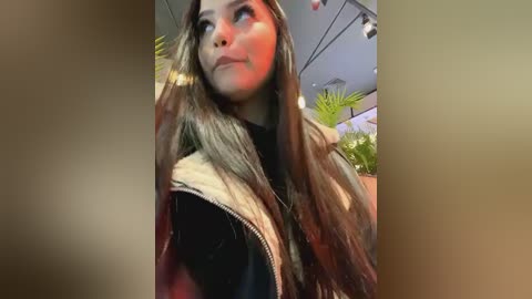 Media: Video of a young woman with long, wavy brown hair, wearing a black jacket, looking contemplative in a modern indoor setting with green plants and overhead lights.