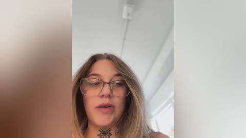 Media: Video of a young woman with fair skin, glasses, and long brown hair, wearing a black floral top. She is indoors with white walls and a power outlet on the ceiling.