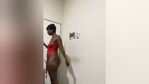 Media: Video of a slender Black woman in a red lace bodysuit, standing in a narrow hallway with white walls and a thermostat, looking away.