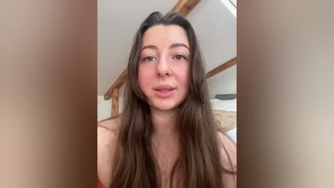 Media: Video of a young, fair-skinned woman with long brown hair, wearing a red top, standing in a bedroom with wooden beams and a bed in the background.