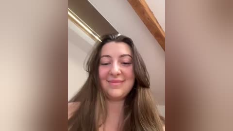 Media: A blurry video of a young woman with long, straight brown hair, fair skin, and a slight smile. She is indoors, possibly in a bathroom, with a wooden ceiling beam visible.