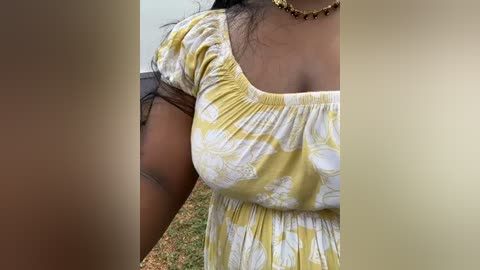 Media: A close-up video of a Black woman with medium brown skin, wearing a yellow floral dress with a rounded neckline, outdoors with a blurred background of grass and a wall.