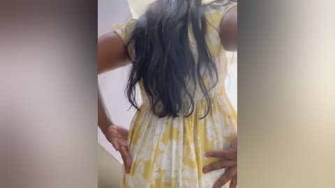 Media: Video of a woman with long, wavy black hair wearing a yellow and white tie-dye dress, standing with hands on hips, viewed from behind in a brightly lit room.