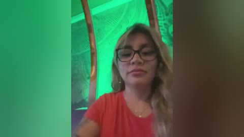 Media: A video of a young woman with long blonde hair and glasses, wearing a red t-shirt, standing in a dimly lit room with greenish lighting and hanging tapestries.