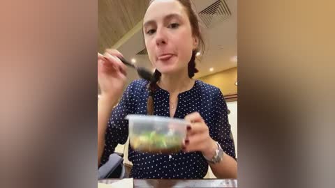 Media: A video of a woman with light skin, brown hair, and fair complexion, eating salad with a spoon, wearing a navy blue polka-dot blouse. The background shows a modern restaurant interior with beige walls and ceiling lights.