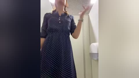 Media: Video of a woman with light skin, wearing a dark blue dress with white polka dots, standing in a bathroom with a white tiled wall and a silver soap dispenser.