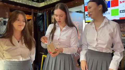 Media: Video of three young Caucasian women in white button-up shirts and gray skirts, examining a bottle in a cozy, dimly-lit bar.