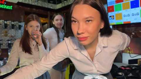 Media: Video of three young women in a bar, one with short dark hair and a light blouse, others with long hair in white shirts, background includes colorful posters and a neon sign.