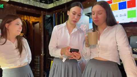 Media: Video of three young women, Caucasian, in casual white blouses and gray skirts, drinking coffee in a cozy, wooden-walled cafe with colorful digital signage.