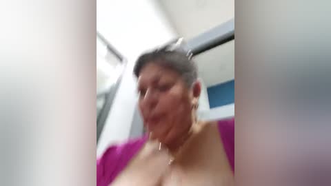 Media: A blurry video of an older woman with short gray hair, wearing a purple top, seen from the chest up, inside a train.