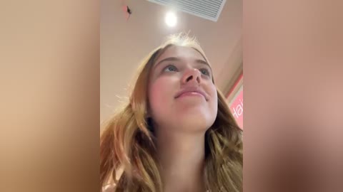 Media: A video of a young Caucasian woman with long, wavy blonde hair, looking upward, with a soft pink ceiling and a vent visible.