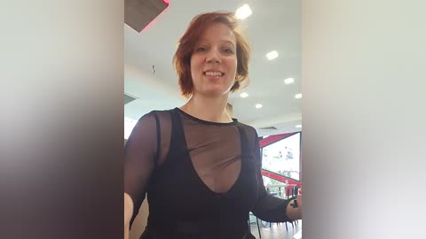 Media: Video of a smiling, fair-skinned woman with short red hair, wearing a black sheer top with a deep V-neckline, standing in a modern, well-lit room with white walls and recessed lighting.