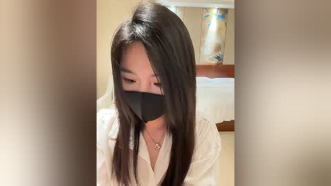 Media: Video of a young Asian woman with long black hair, wearing a black face mask and white robe, sitting indoors with blurred background featuring a bed and abstract art.