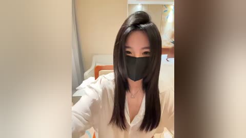 Media: A video of an East Asian woman with long black hair, wearing a black face mask, a white button-up shirt, and sitting on a bed in a room with beige walls and a wooden nightstand.