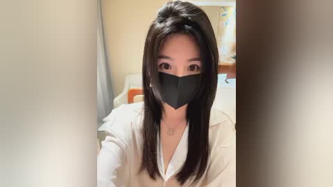 Media: A video of an Asian woman with long black hair, wearing a black mask, white shirt, and a gold necklace, taken indoors in a dimly lit room.