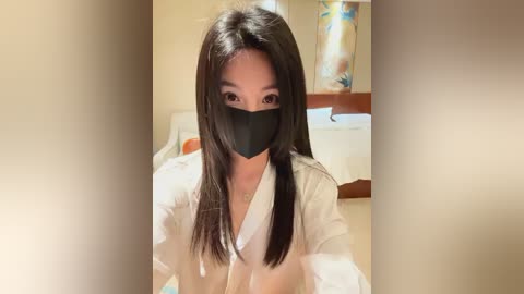 Media: A video of an Asian woman with long, straight black hair, wearing a white shirt, black face mask, and a gold necklace, in a softly lit room with beige walls and a floral painting.