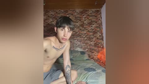 Media: Video of a shirtless young Asian man with short dark hair, sitting on a bed with green bedding. He has a tattoo on his chest and arm. Brick wall background, dim lighting.