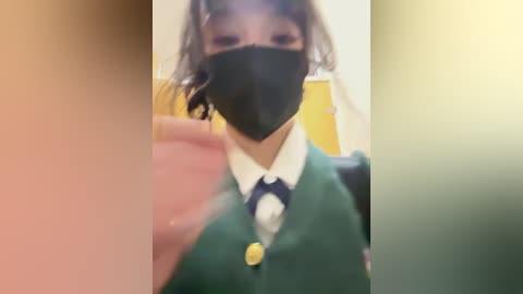 Media: A video of a person wearing a green vest, white shirt, and blue tie with a yellow button, black face mask, and black hair. The image is slightly blurred and has a soft focus.