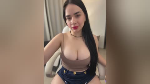 Media: Video of a Latina woman with long black hair, wearing a beige crop top revealing large breasts and high-waisted denim shorts, sitting on a white chair in a beige room.