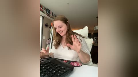 Media: A candid video of a fair-skinned woman with long brown hair, wearing a white top, sitting at a computer desk, smiling and waving, surrounded by cluttered shelves and a white chair.