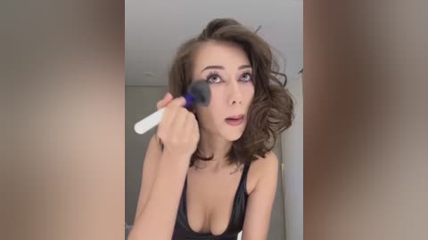 Media: Video of a woman with wavy brown hair applying makeup with a white brush, wearing a black lace bra.