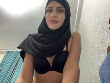 Media: A video of a woman with medium skin tone, wearing a black hijab and black bra, standing indoors against a textured white wall.