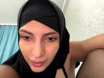 Media: A close-up video of a woman with fair skin, brown eyes, and dark brown hair partially covered by a black hijab. She has a neutral expression and is wearing a black sleeveless top. The background features a white wall with a light pattern and a turquoise curtain.