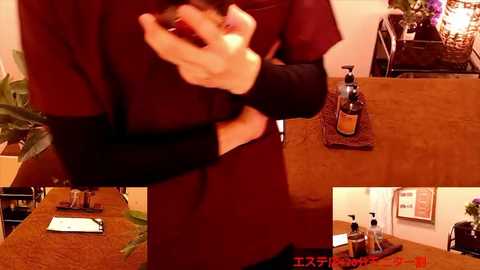 Media: Video of a person in a dark maroon shirt, hand clasped over their chest, standing in a warmly lit office with a wooden desk and potted plants, bottles of hand sanitizer visible.