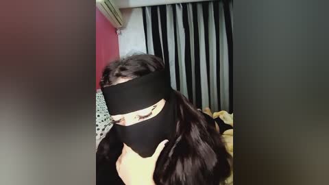 Media: A video of a person with dark hair and a black face mask, standing in a room with pink walls, a bed, and a window with black curtains.