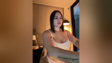 Media: Video of a young woman with medium skin tone, straight black hair, wearing a white spaghetti-strap tank top and pink underwear. She sits in a dimly lit room, with a black lamp on a wooden table in the background.