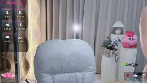 Media: A video captures a cozy, light-filled room with a plush, gray recliner, a white lamp, a pink plush toy, and sheer curtains. A video game screen displays a gameplay scene in the background.