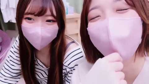 Media: Video of two young Asian women with long brown hair, wearing pink face masks, in striped and white tops, indoors with blurred background.