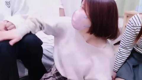 Media: Video of a young Asian woman with short brown hair, wearing a white sweater, black skirt, and pink mask, sitting on a bed with a white duvet.