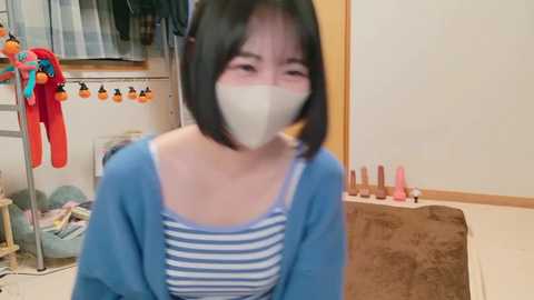 Media: Video of an East Asian woman with short black hair, wearing a blue cardigan over a striped top, mask, and glasses, in a cluttered room with hanging clothes and sex toys.