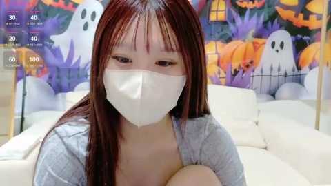 Media: Video of a young Asian woman with long red hair, wearing a white mask, light gray sweater, and white background featuring Halloween decor with pumpkins and ghosts.