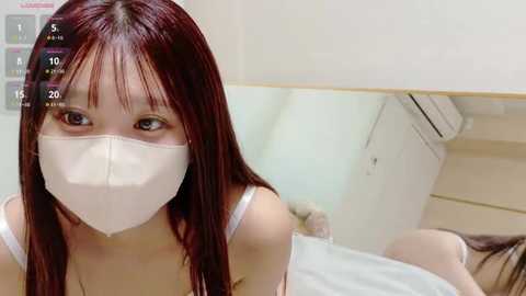 Media: A video of a young East Asian woman with long, straight, dark hair, wearing a white surgical mask and a white tank top, sitting in a hospital room with a white bed and air conditioning unit in the background.