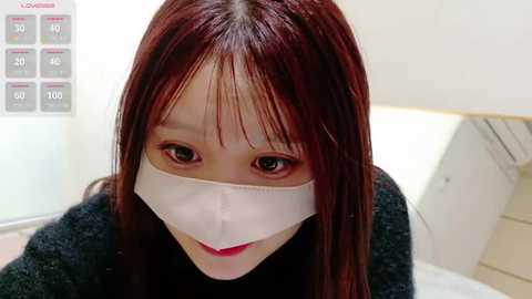 Media: Video of an Asian woman with long, straight, dark brown hair, wearing a white surgical mask, black top, and a subtle smile, indoors with light-colored walls and a white door.