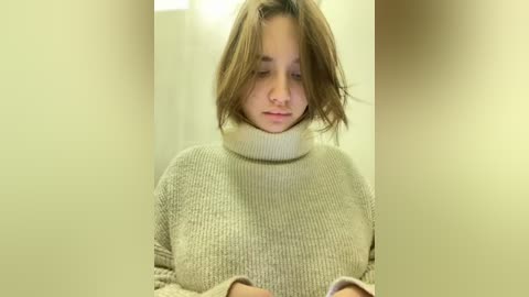 Media: Video of a young woman with light skin and brown hair, wearing a beige knitted turtleneck sweater, looking down with a neutral expression in a dimly lit room with off-white walls.