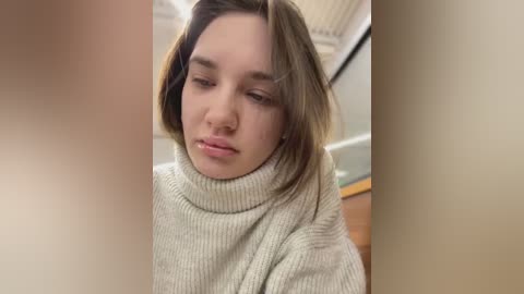 Media: A video of a young woman with shoulder-length, light brown hair, wearing a cream-colored turtleneck sweater, looking down thoughtfully. The background shows a blurred indoor setting with beige walls and a wooden table.