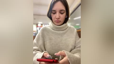 Media: Video of a young woman with shoulder-length dark hair, wearing a beige turtleneck sweater, intently looking at a red smartphone in a brightly lit, indoor setting with blurred background.