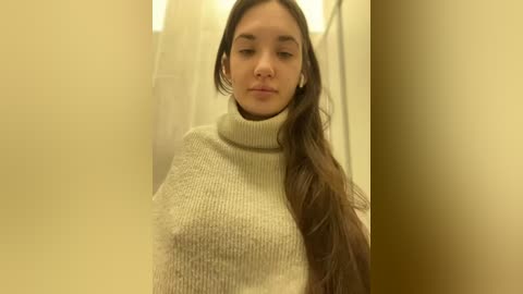 Media: Video of a young woman with long brown hair, wearing a cream-colored turtleneck sweater, standing against a blurred, beige background. She has a neutral expression.