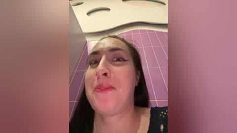 Media: Video of a young woman with fair skin and straight dark hair, wearing pink lipstick and a black lace dress, standing in a purple-tiled bathroom with wavy white ceiling panels.