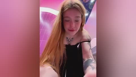 Media: Video of a young woman with long, straight blonde hair, wearing a black off-shoulder top, with tattoos on her chest and arms, sitting in a pink-tiled bathroom.