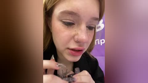 Media: Video of a young woman with fair skin and light brown hair, wearing a black jacket, meticulously applying makeup. She has a septum piercing and is focused on her eyelashes. Background features a purple wall with partial text.