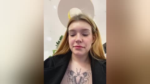 Media: Video of a young, fair-skinned woman with long, straight blonde hair, wearing a black jacket, and a prominent butterfly tattoo on her chest. She stands indoors with white balloons and a green plant in the background.