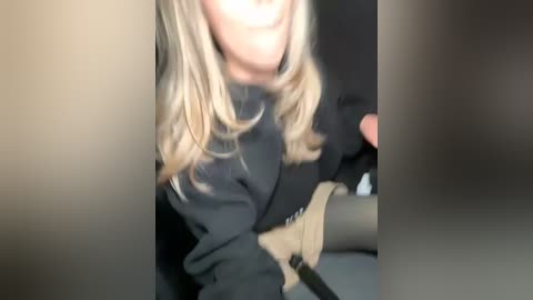 Media: Video of a blonde woman with shoulder-length hair, wearing a black hoodie, seated with a beige leg brace on her right leg, captured from a low angle.