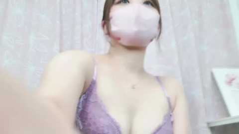 Media: Video of a young Asian woman with light skin, wearing a lavender lace bra and pink face mask. She has a slim physique with small breasts and brown hair tied back. The background features a light pink floral curtain.