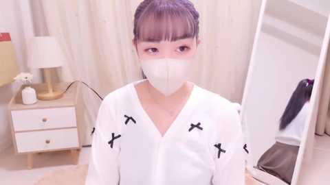 Media: Video of an East Asian woman with pale skin, wearing a white mask, white V-neck shirt with black crosses, and a white headband, sitting in a softly lit, minimalist bedroom with white curtains and a wooden nightstand.