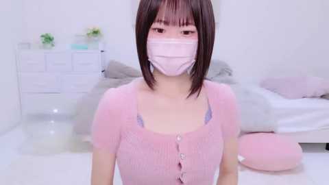 Media: Video of an Asian woman with straight brown hair, wearing a pink face mask, pink cardigan, and pastel-colored bra, in a minimalistic white bedroom with a bed, dresser, and floral decor.