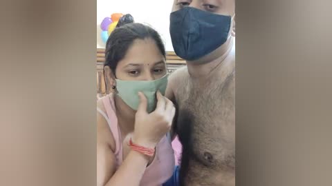 Media: Video of a South Asian woman in a pink tank top and green mask, holding a man's hand, with a hairy, shirtless man wearing a blue mask in the background.